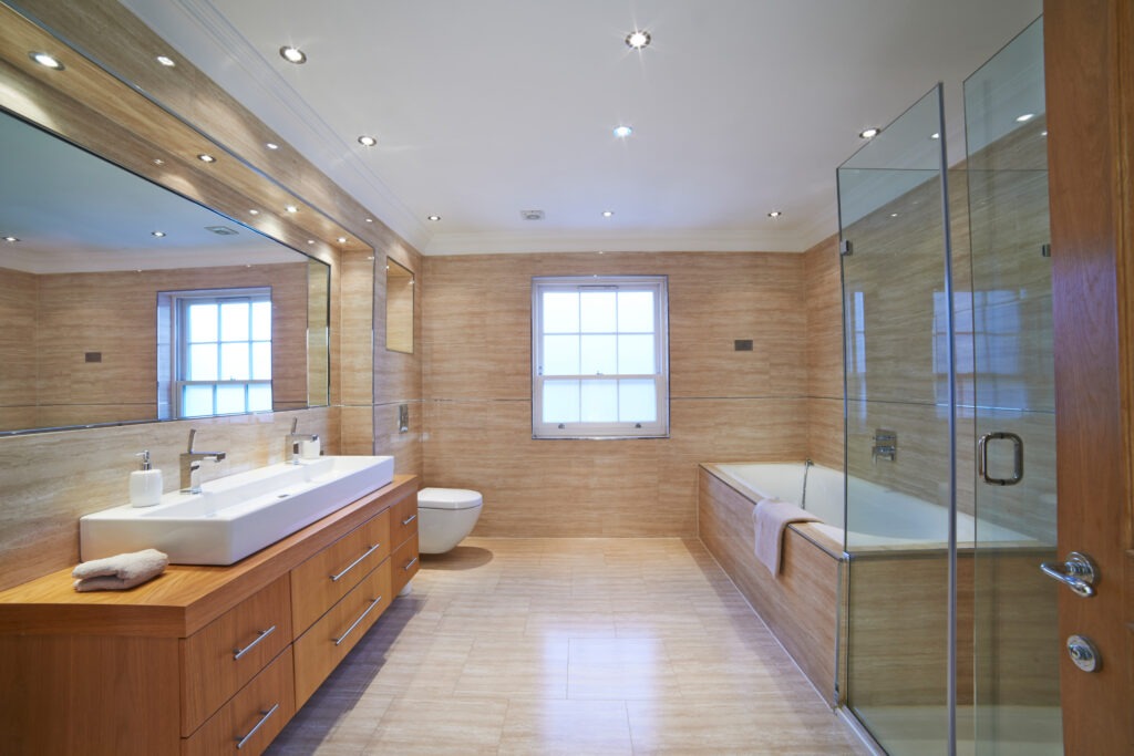 Modern bathroom featuring a double sink vanity, glass shower enclosure, and bathtub. Natural light floods the space through a window, enhancing the warm wood tones.